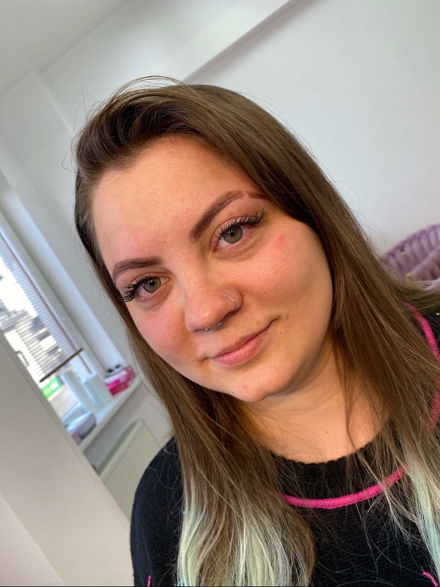 Portret kobiety z delikatnie podkreślonymi brwiami, świeżo po zabiegu, z widocznym zaczerwienieniem skóry wokół brwi i na policzku. Widoczny kolczyk w nosie i przedłużone rzęsy.