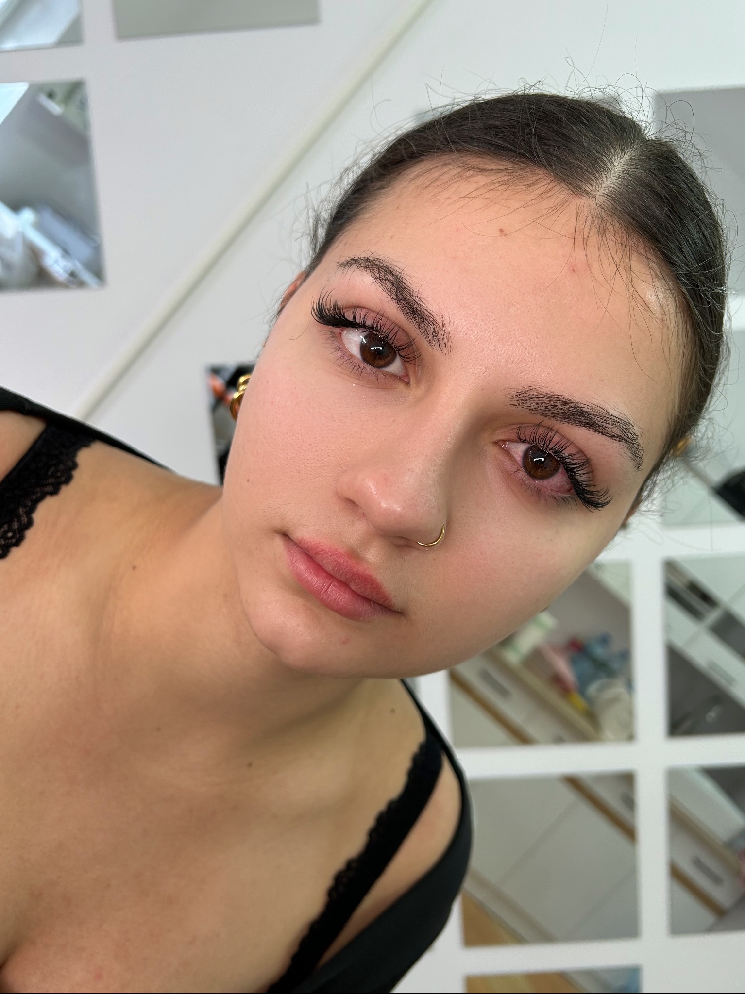 Portret kobiety z brązowymi oczami i świeżo przedłużonymi rzęsami, widoczny złoty kolczyk w uchu i kolczyk w nosie, na twarzy widoczne delikatne zaczerwienienia.