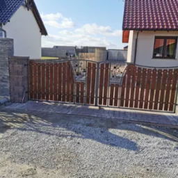 Dwuskrzydłowa brama wjazdowa z drewnianymi wypełnieniami i metalowymi zdobieniami, osadzona na murowanych słupkach z kostki, widok na posesję z szarego żwiru i dwa domy w tle.