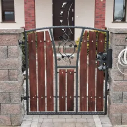 Kuta furtka z drewnianymi wypełnieniami, zdobiona motywem roślinnym, osadzona między murowanymi słupkami z bloków betonowych, z widocznym przewodem na słupku.