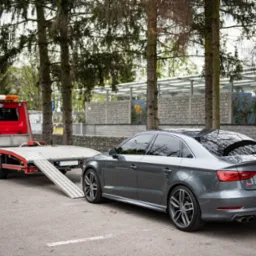 Szara limuzyna Audi S3 na rampie czerwonej lawety z otwartą platformą, zaparkowana na parkingu z drzewami w tle.