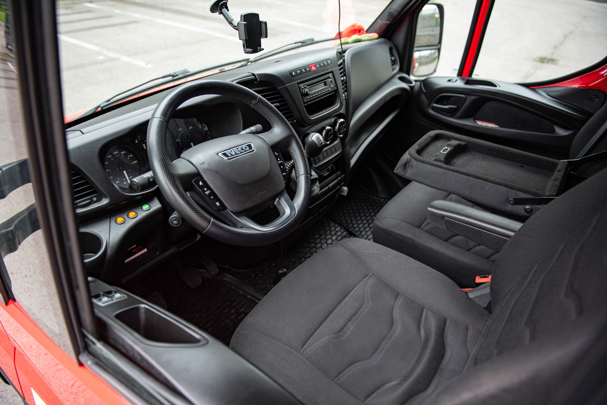 Wnętrze kabiny czerwonego busa marki Iveco, widok z otwartych drzwi na deskę rozdzielczą, kierownicę z logo, czarne fotele i gumowe dywaniki.
