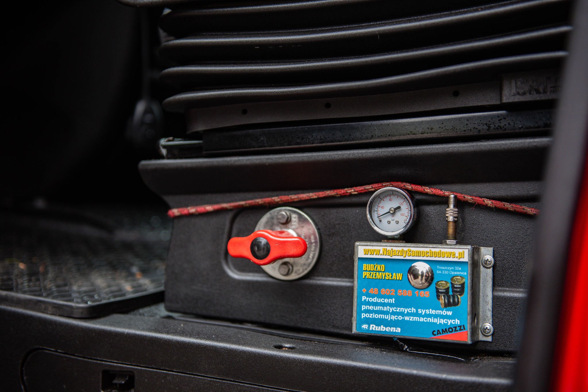 Mechanizm pneumatycznego zawieszenia z manometrem i zaworem odcinającym, zamontowany pod fotelem w kabinie samochodu dostawczego, widoczna tabliczka informacyjna producenta systemów pneumatycznych.
