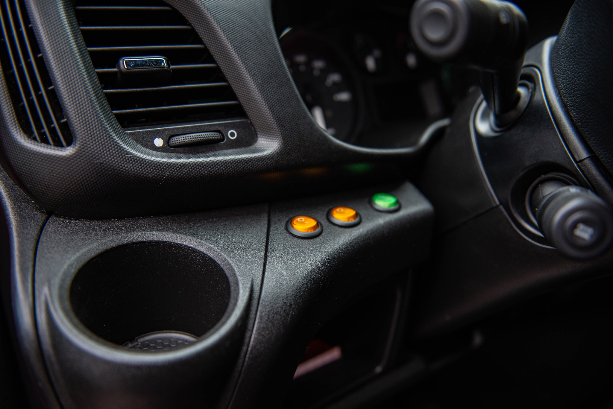 Wnętrze kabiny busa: deska rozdzielcza z kratką wentylacyjną, dwoma pomarańczowymi i jednym zielonym przyciskiem oraz uchwytem na napoje, widok z bliska.