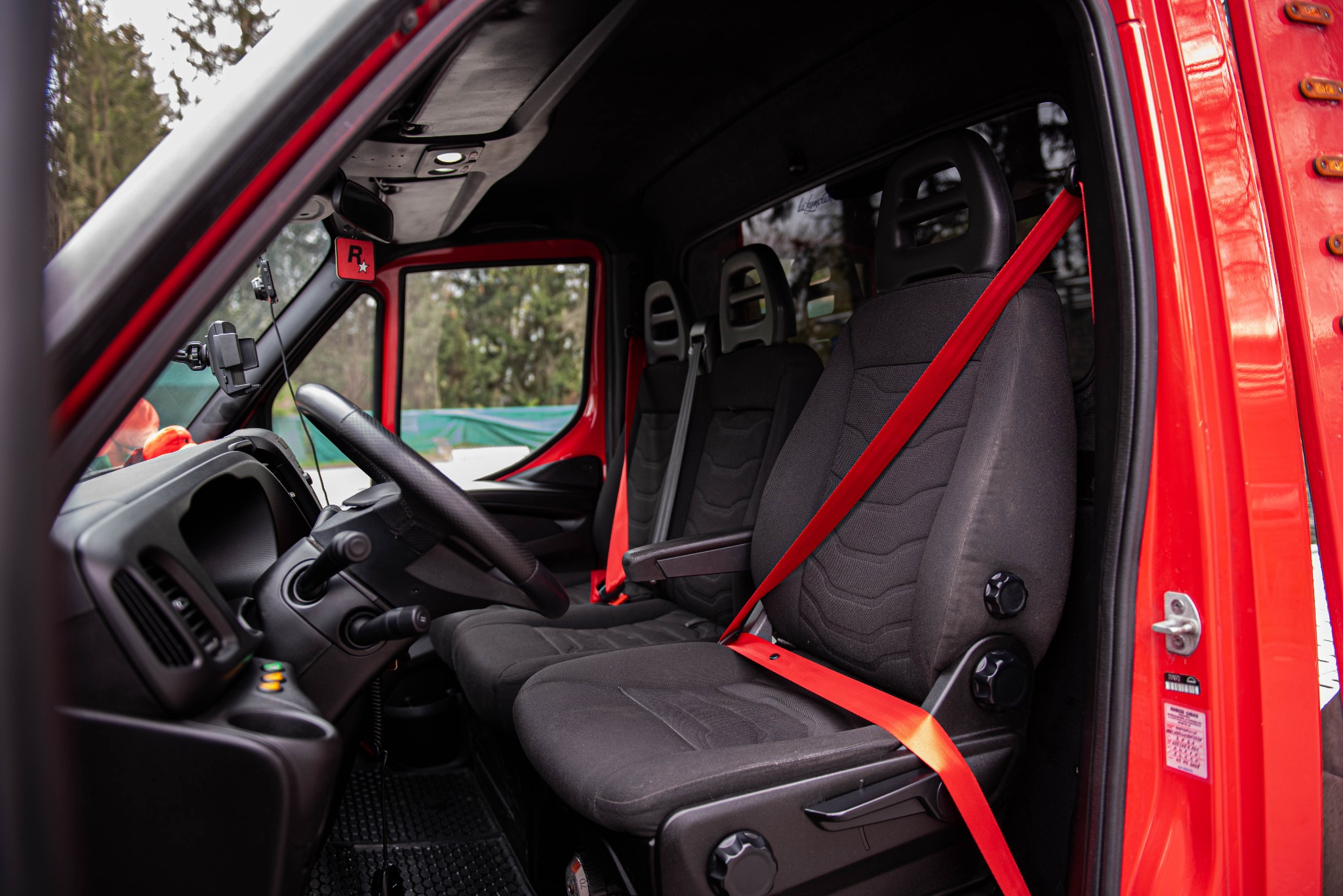 Wnętrze czerwonego busa z czarnymi fotelami i czerwonymi pasami bezpieczeństwa, widok z zewnątrz przez otwarte drzwi kierowcy, z uchwytem na telefon na desce rozdzielczej.