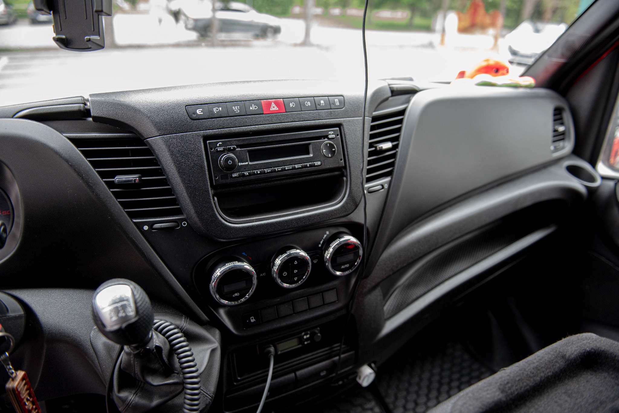 Wnętrze kabiny samochodu dostawczego z widoczną deską rozdzielczą, radiem, panelem klimatyzacji i dźwignią zmiany biegów. Na desce znajduje się rząd przycisków funkcyjnych. W tle widoczne...