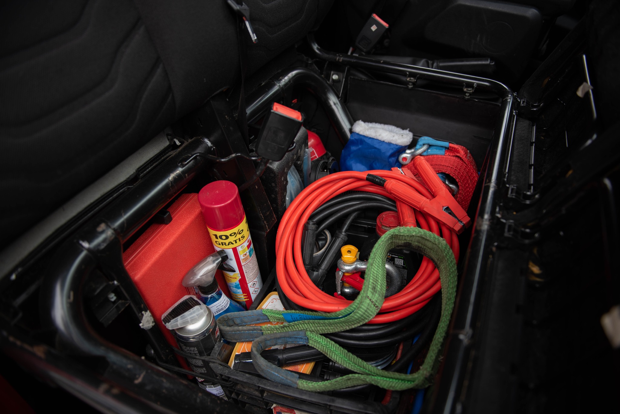 Schowek w samochodzie wypełniony akcesoriami: kable rozruchowe, lina holownicza, preparaty w sprayu, zestaw narzędzi, pasy transportowe.