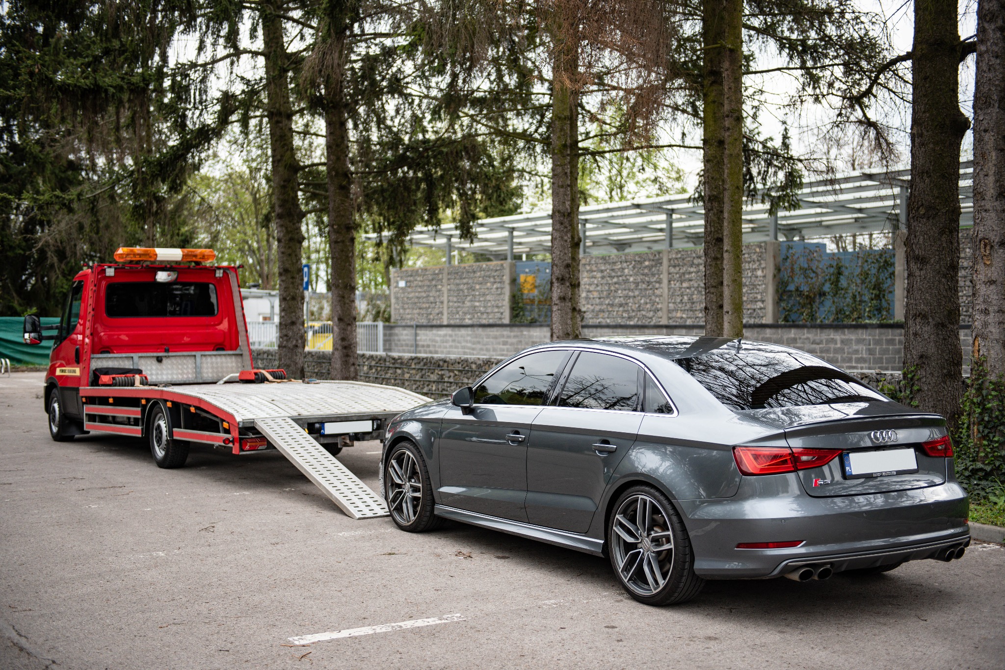 Szara limuzyna Audi S3 na rampie czerwonej lawety z otwartą platformą, zaparkowana na parkingu z drzewami w tle.