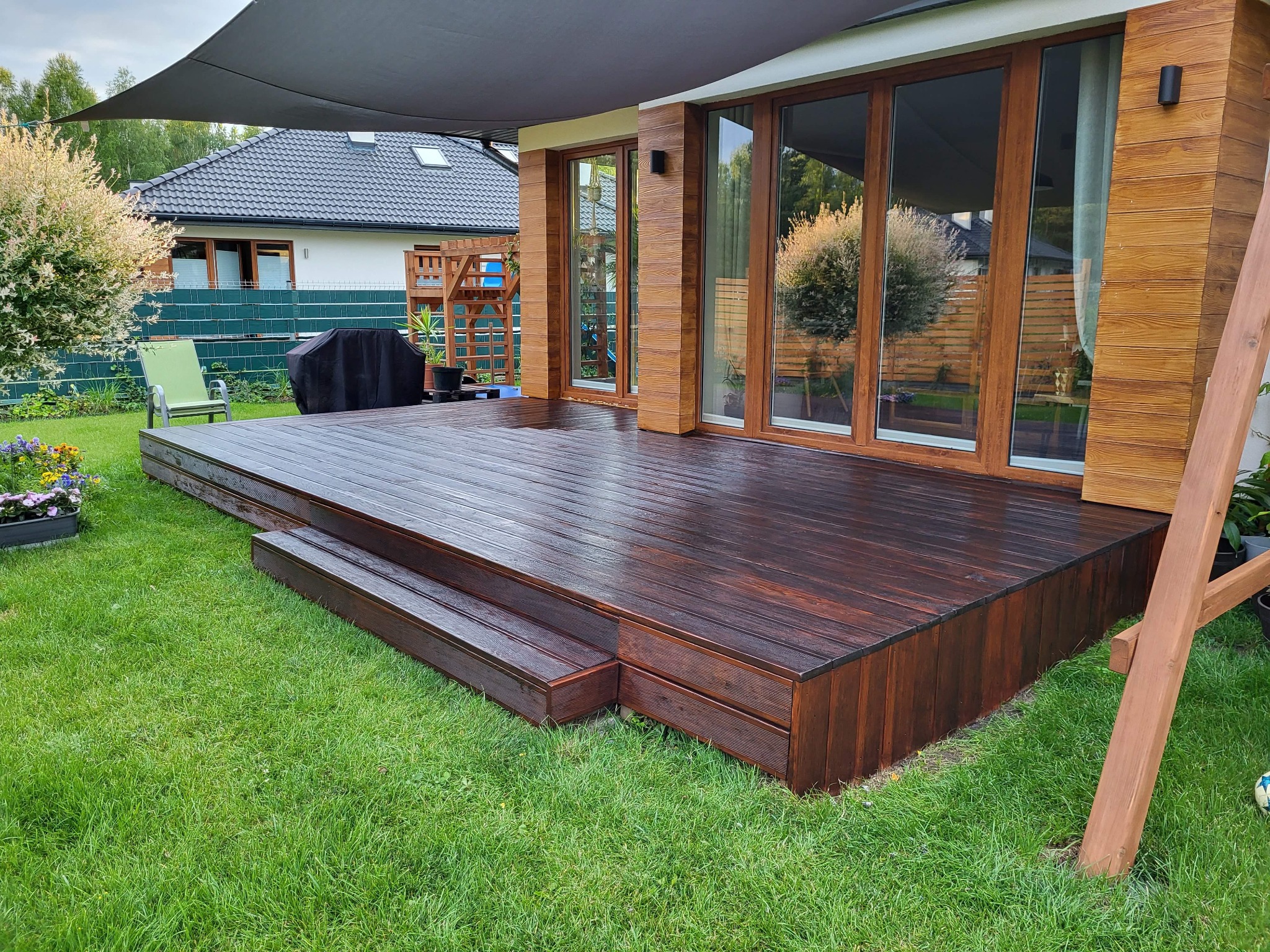 Drewniany taras z dwoma stopniami, świeżo po impregnacji, zadaszony szarą markizą w Kałuszynie, z widokiem na dom i ogród.