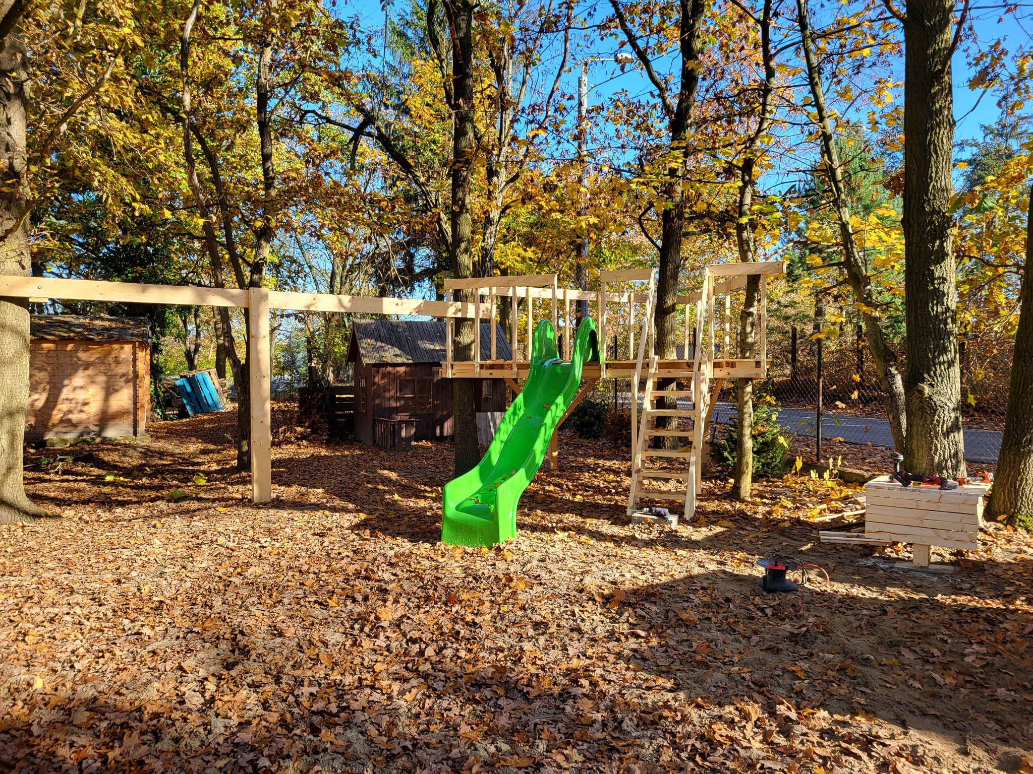 W trakcie budowy drewniany plac zabaw z zieloną zjeżdżalnią, drabinką i domkiem na drzewie, otoczony jesiennymi drzewami i opadłymi liśćmi. W tle widoczne narzędzia i materiały budowlane.