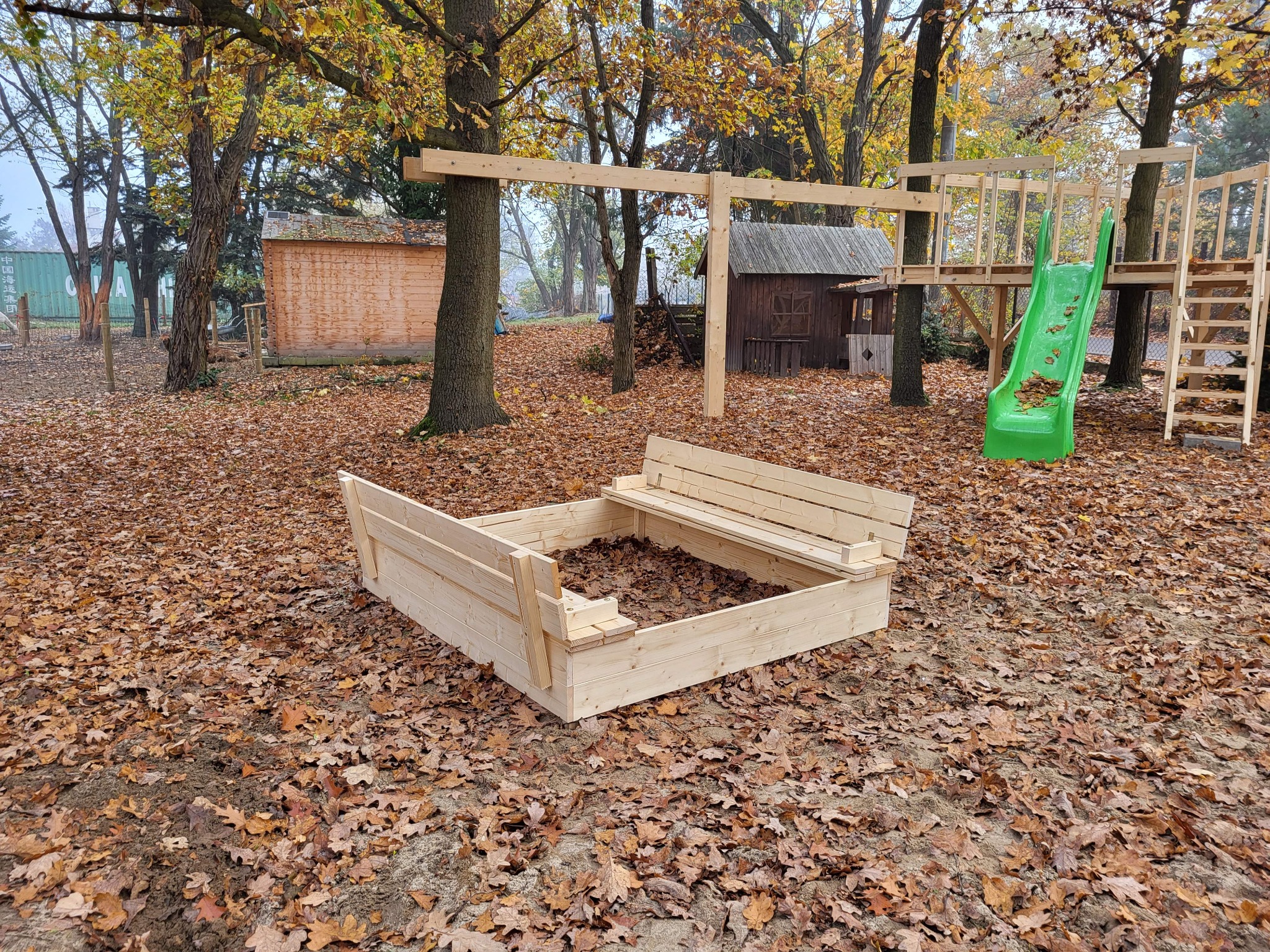 Plac zabaw z piaskownicą z siedziskami i drewnianą konstrukcją ze zjeżdżalnią, otoczony opadłymi liśćmi.