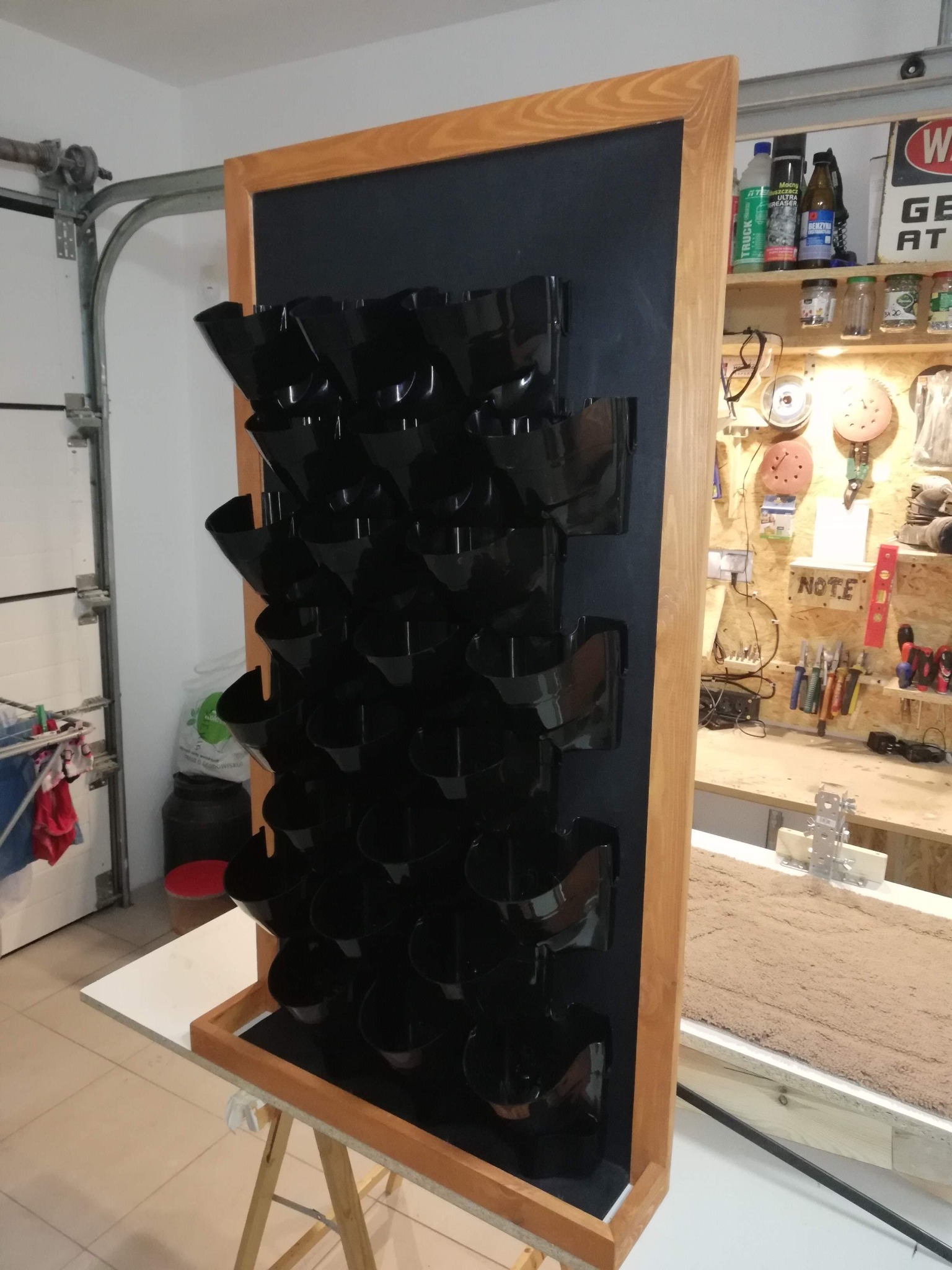 Drewniana rama z czarnymi doniczkami na ścianę w warsztacie, widoczne narzędzia i akcesoria na półkach w tle.