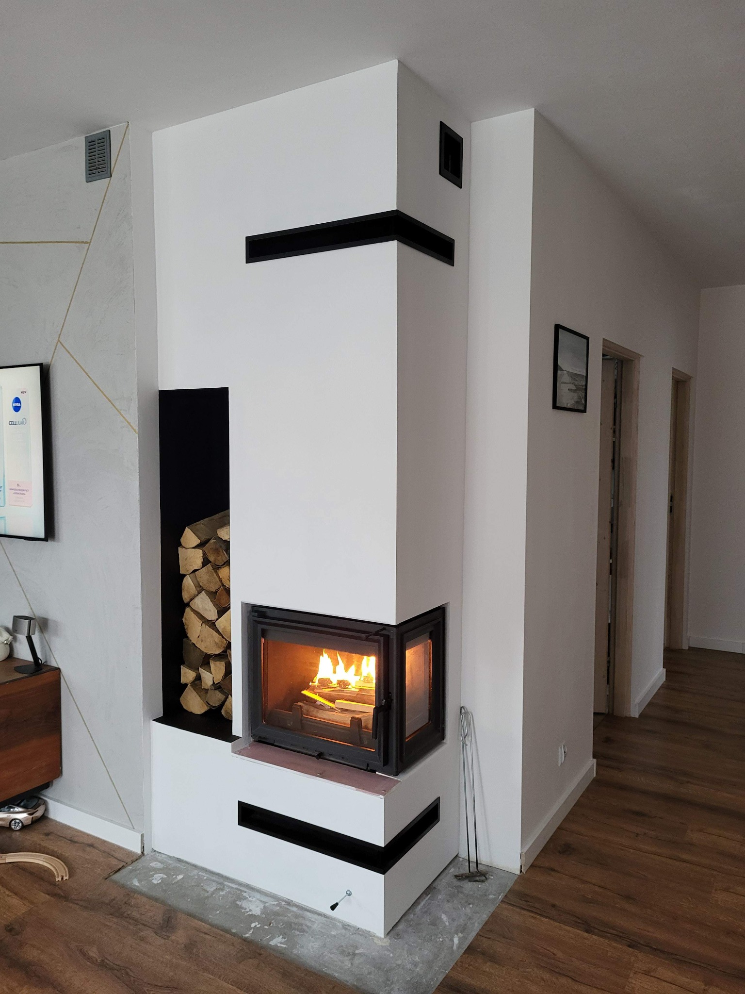 Nowoczesny narożny kominek z czarnymi akcentami, płomieniami i zapasem drewna, widok na drewnianą podłogę i częściowo wykończoną wylewkę, w tle drzwi i dekoracyjna ściana.