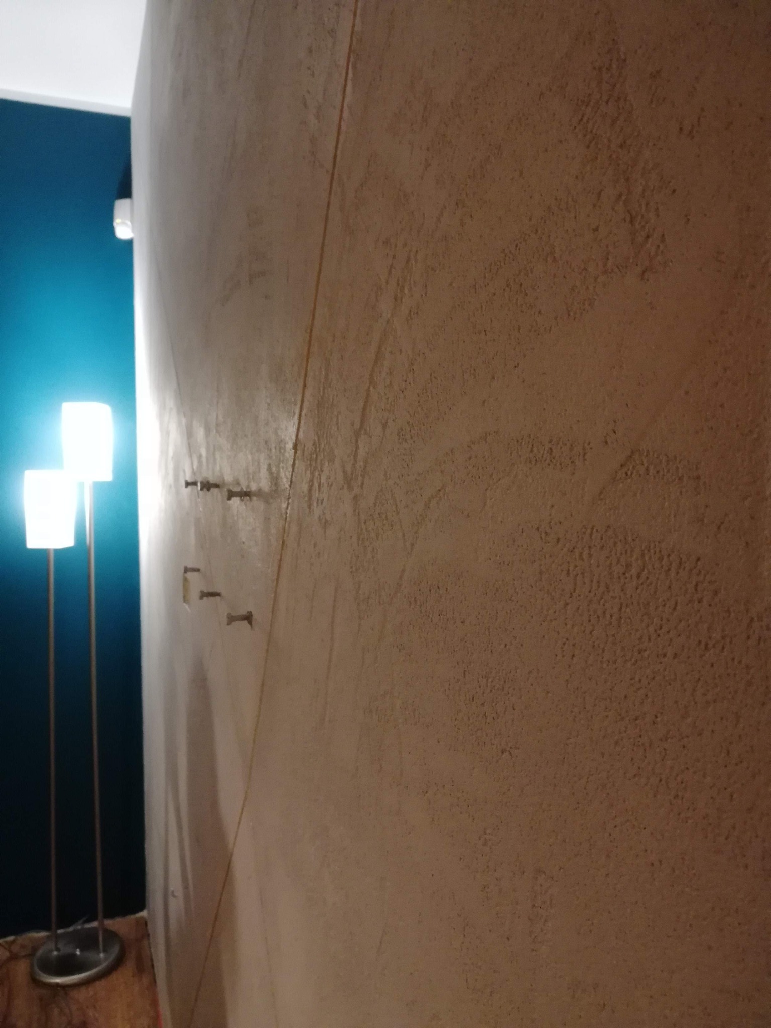 Beżowa ściana z fakturą, oświetlona przez dwie lampy stojące, z widocznymi śladami po mocowaniach.