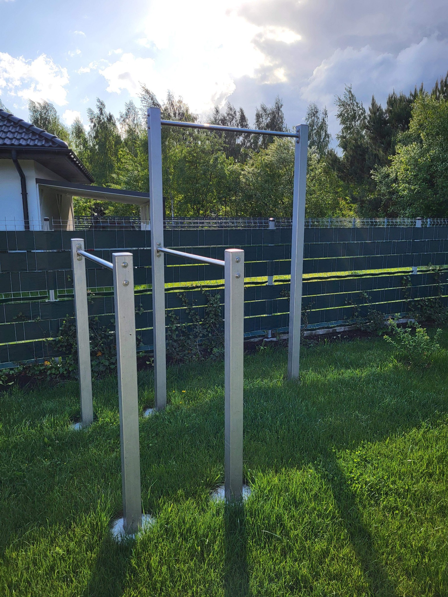 Metalowa konstrukcja do ćwiczeń na świeżym powietrzu, z drążkami do podciągania i poręczami, umieszczona na trawniku przed zielonym ogrodzeniem i budynkiem w tle.