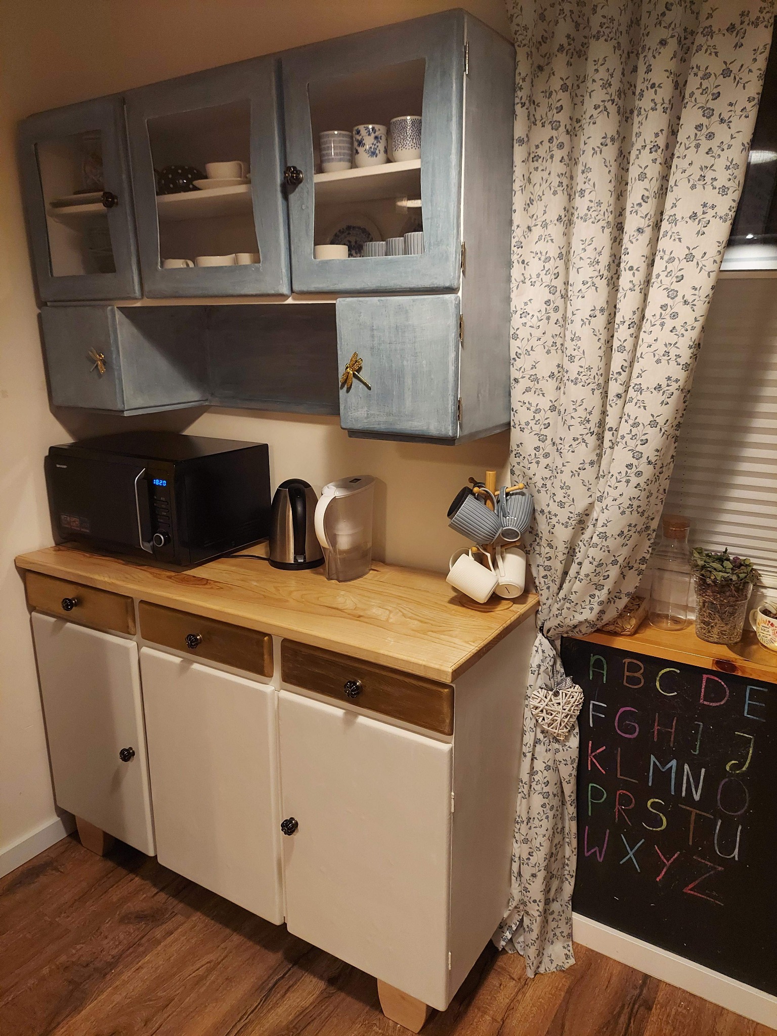 Kuchenna zabudowa w stylu vintage: błękitne szafki wiszące z przeszklonymi drzwiczkami, białe szafki dolne z drewnianym blatem, mikrofalówka, czajnik, wieszak na kubki, zasłona w kwiaty i tablica...