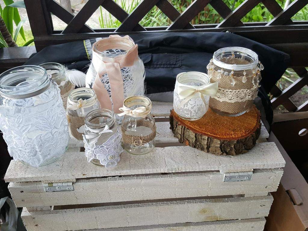 Dekoracyjne słoiki z koronką, jutą i wstążkami, ustawione na pomalowanej na biało drewnianej skrzynce i drewnianym plastrze, tworzące ozdobę w stylu rustykalnym.
