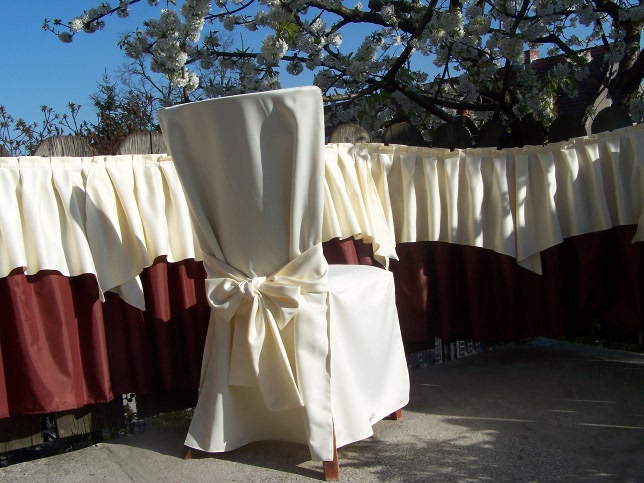 Krzesło udekorowane kremowym pokrowcem z satynową kokardą na tle białego obrusu z falbanami i bordową podszewką, na zewnątrz w słoneczny dzień.