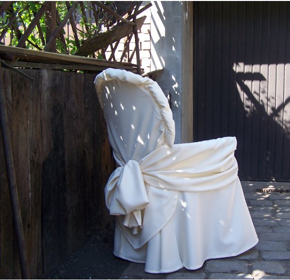 Krzesło z kremowym pokrowcem i ozdobnym wiązaniem, sfotografowane na zewnątrz, w tle drewniany płot i brama garażowa.