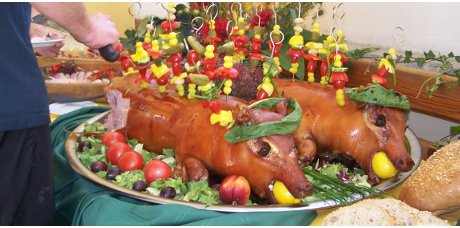 Dwa pieczone prosięta na srebrnej tacy, udekorowane warzywami na szpikulcach i owocami, z cytryną w pyszczku, prezentowane na stole z zielonym obrusem.