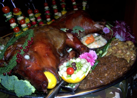 Pieczone prosię z cytryną w pysku, udekorowane owocami i warzywnymi szaszłykami, podane na srebrnej tacy z różnymi rodzajami sałatek i surówek.