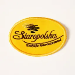 Ż&oacute;łta, owalna naszywka z wyhaftowanym czarnym napisem 'Staropolska. Tradycja Nowoczesności' oraz symbolem graficznym powyżej napisu, leżąca na białym tle.