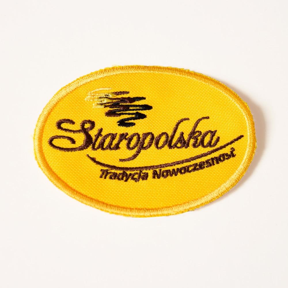 Żółta, owalna naszywka z wyhaftowanym czarnym napisem 'Staropolska. Tradycja Nowoczesności' oraz symbolem graficznym powyżej napisu, leżąca na białym tle.