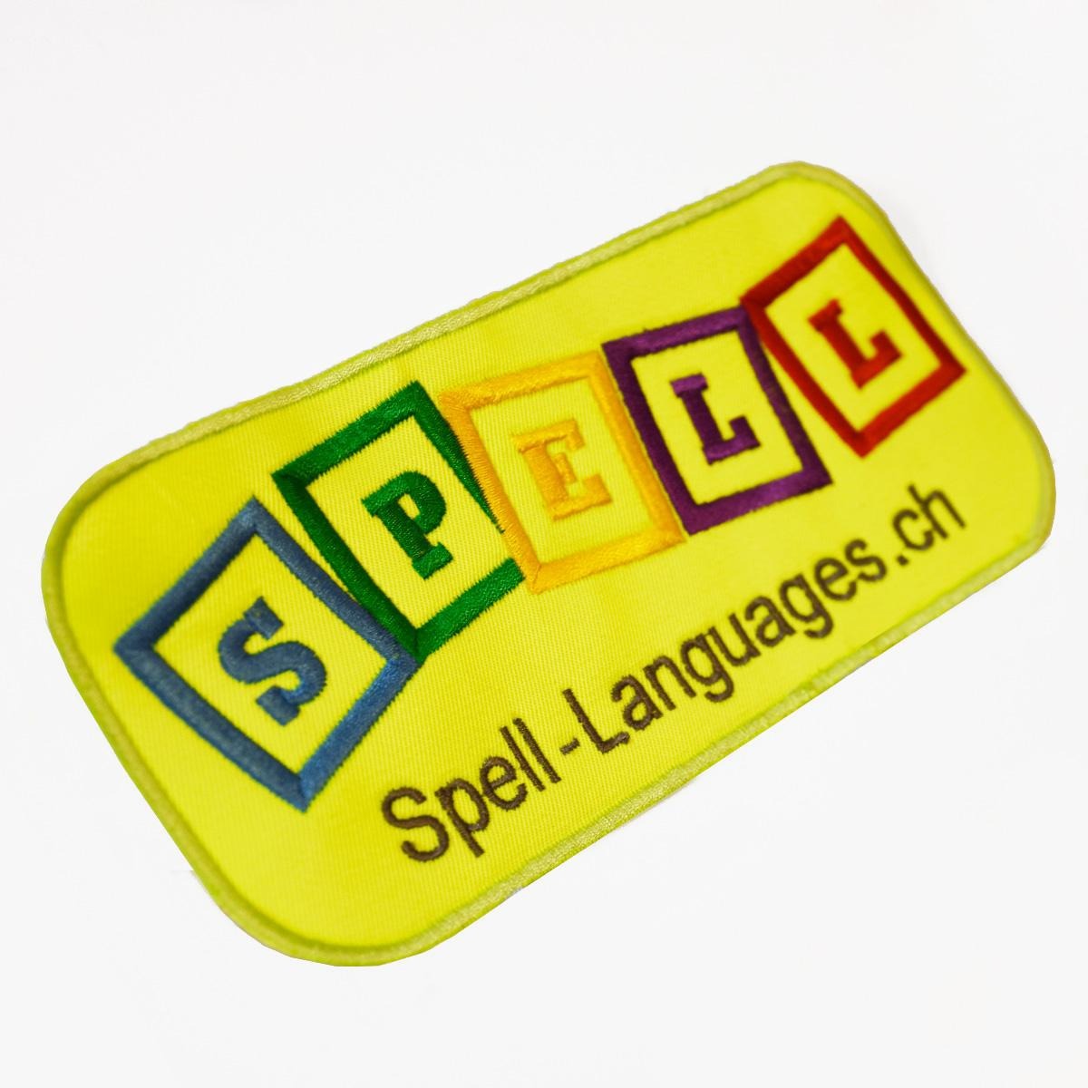 Żółta naszywka z wyhaftowanym kolorowym napisem 'SPELL' w kwadratowych ramkach i adresem strony internetowej Spell-Languages.ch