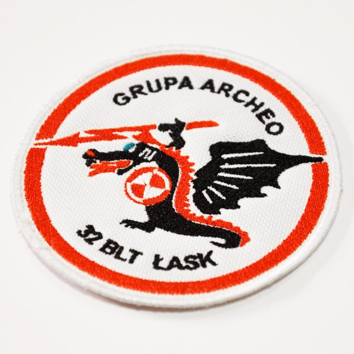 Okrągła naszywka z wyhaftowanym czarnym smokiem z czerwonymi akcentami i napisem 'GRUPA ARCHEO' oraz '32 BLT ŁASK' na białym tle z czerwoną obwódką.