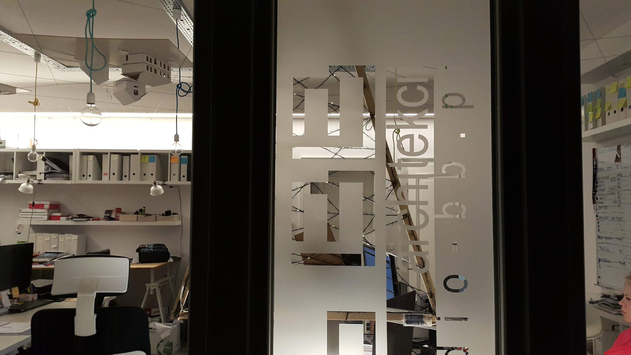 Szklane drzwi biura z wyciętym logo firmy, widok na wnętrze z biurkami, regałami z segregatorami i wiszącymi modelami domów.