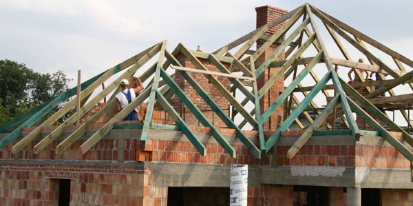 Wznoszenie drewnianej konstrukcji dachu na murowanym domu z cegły, widoczni pracownicy w trakcie montażu elementów więźby, komin z cegły w tle.