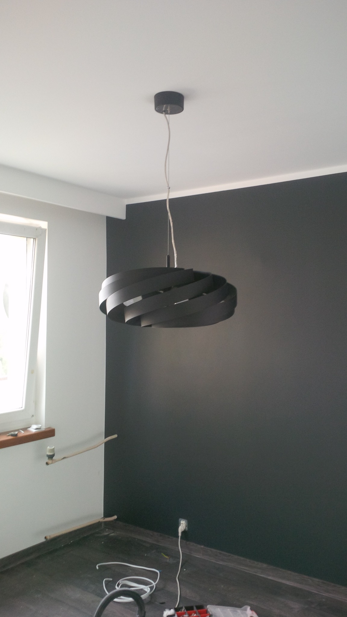 Nowoczesna, spiralna lampa wisząca w kolorze czarnym, zamontowana w pokoju z pomalowaną na ciemny kolor ścianą, widoczne przewody i narzędzia na podłodze wskazujące na prace wykończeniowe.