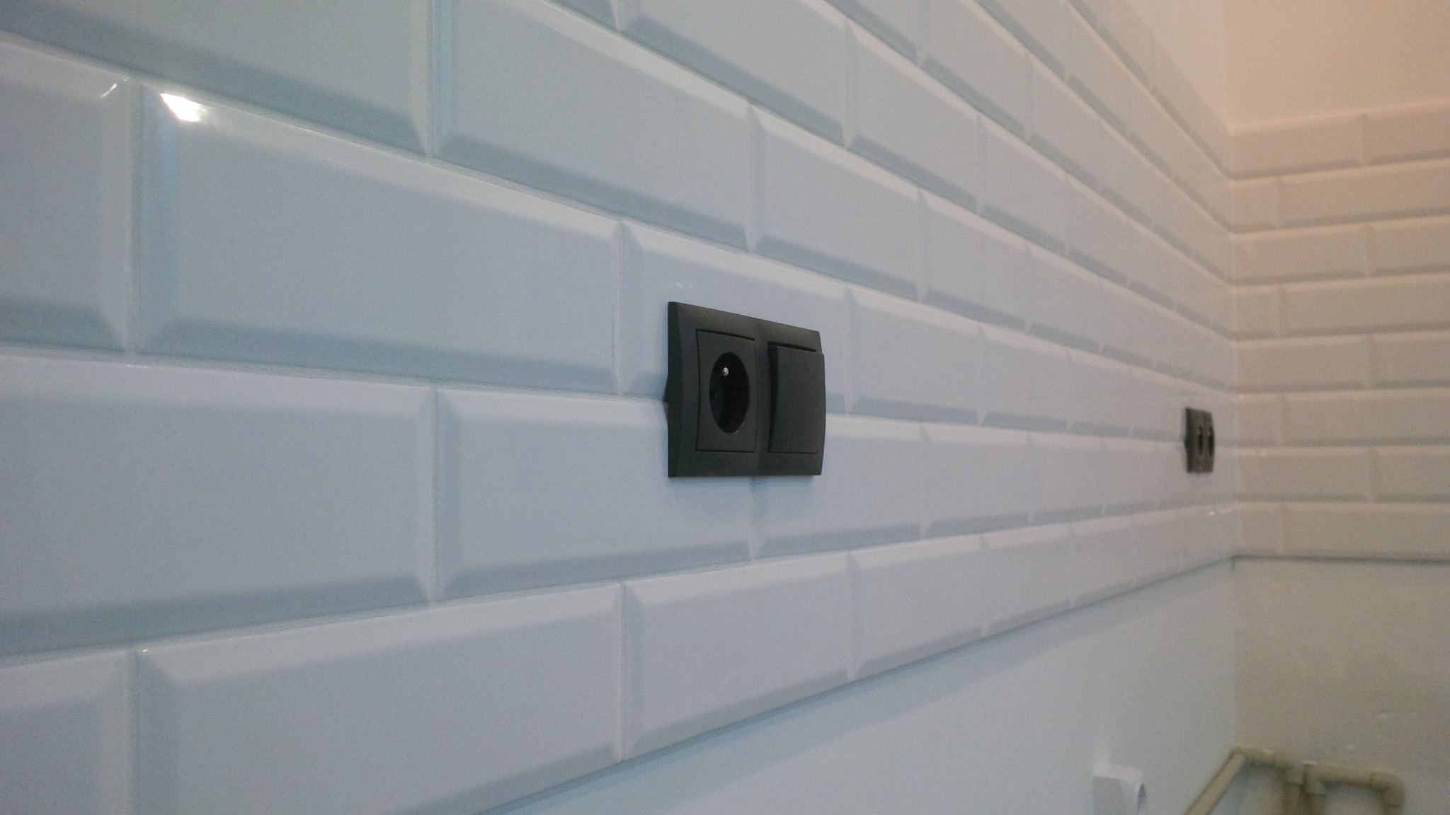 Białe, błyszczące płytki ułożone w cegiełkę z zamontowanym czarnym gniazdkiem elektrycznym i włącznikiem światła.