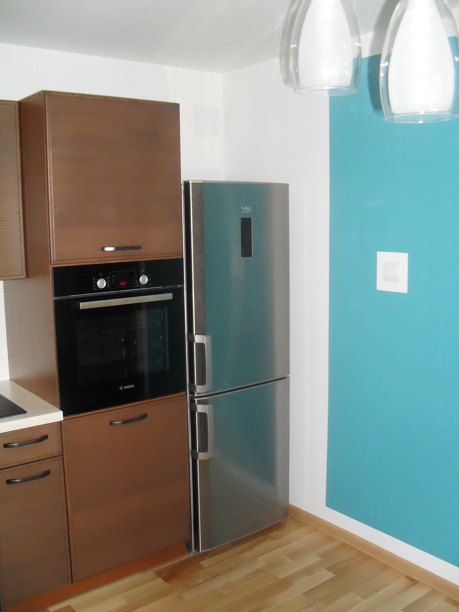 Nowoczesna kuchnia z brązowymi szafkami, czarnym piekarnikiem Bosch i srebrną lodówką Beko na tle turkusowej ściany.