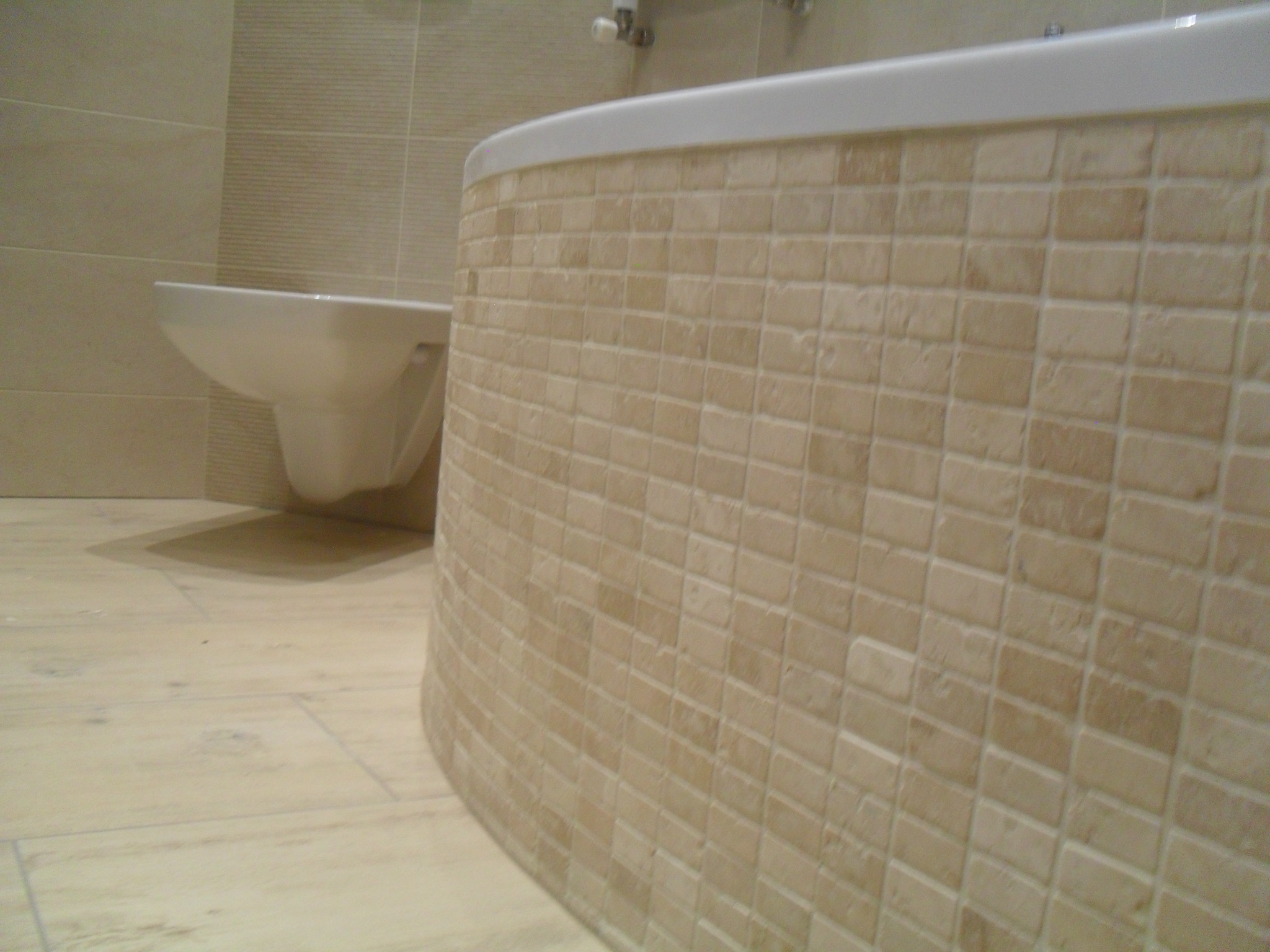 Obudowa wanny wyłożona beżową mozaiką, widoczna toaleta wisząca i podłoga imitująca drewno.