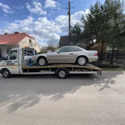 Laweta z beżowym Mercedesem SL na platformie, zaparkowana na ulicy przed domem z czerwonym dachem. Na pojeździe napis 'Pomoc Drogowa' oraz numer telefonu.