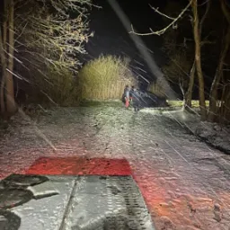 Pomoc drogowa w zimowych warunkach: holowanie pojazdu na zaśnieżonej drodze w nocy, oświetlone reflektorem, widoczna platforma lawety z czerwonymi światłami.