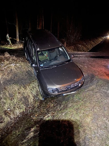 Brązowy SUV Dacia Duster częściowo w rowie przy leśnej drodze, holowany przez osobę z liną, nocne oświetlenie, widoczne światła nadjeżdżającego pojazdu.