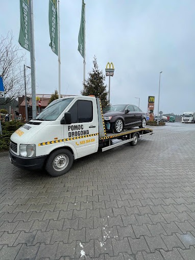 Laweta pomocy drogowej z załadowanym brązowym samochodem osobowym na parkingu z widocznymi w tle znakami McDonald's i innymi pojazdami.