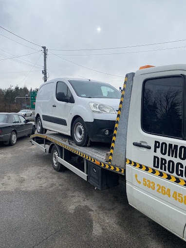 Biały samochód dostawczy przewożony na lawecie pomocy drogowej z numerem telefonu, na tle szarego asfaltu i pochmurnego nieba.