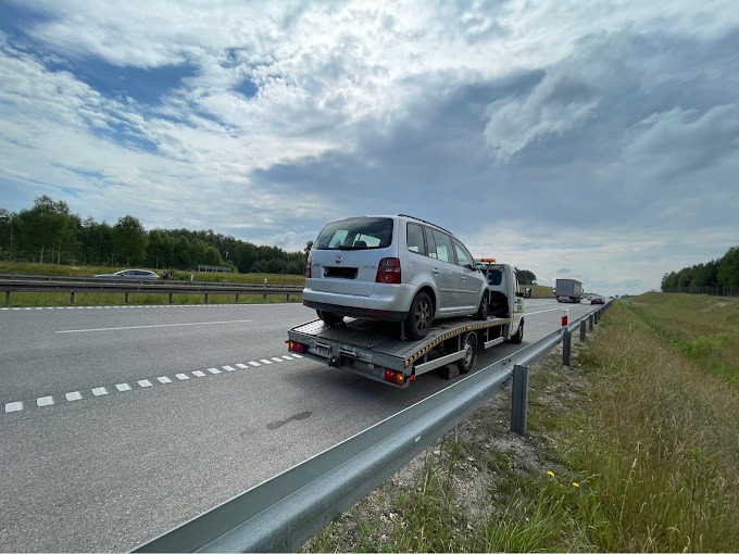 Srebrny samochód osobowy typu kombi przewożony na lawecie na autostradzie w słoneczny dzień.
