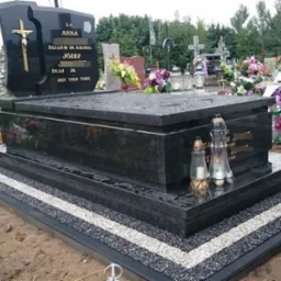 Czarny, granitowy nagrobek z krzyżem i inskrypcjami, otoczony dekoracyjną obwódką z kruszywa w dwóch kolorach, z zapalonymi zniczami i kwiatami w tle.