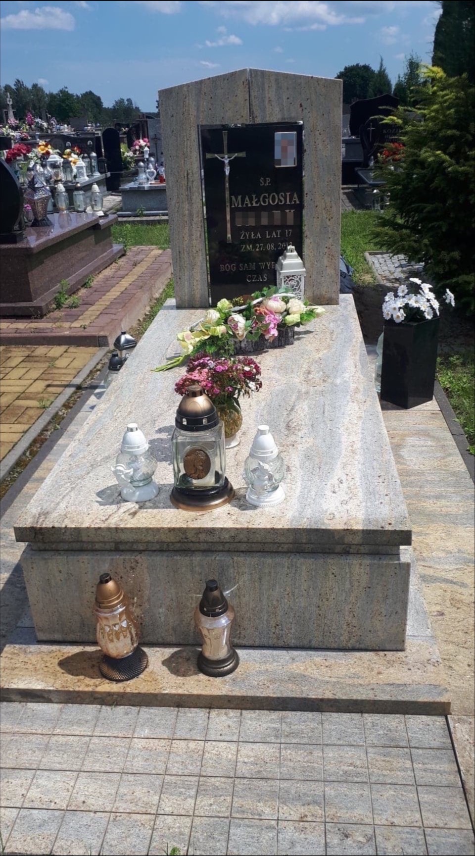 Granitowy pomnik z czarną tablicą nagrobną, ozdobiony kwiatami i lampionami, na cmentarzu w słoneczny dzień.