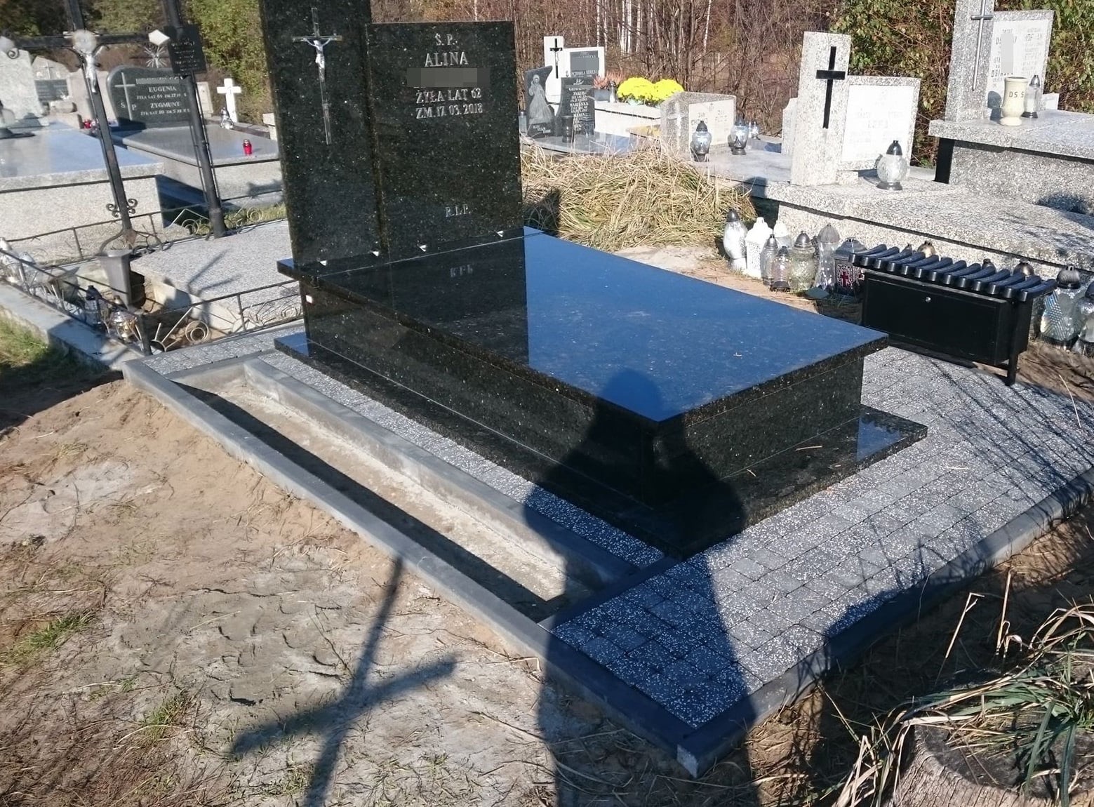 Błyszczący, czarny pomnik z granitu z wygrawerowanym krzyżem i inskrypcją, otoczony ramą z szarej kostki brukowej na cmentarzu.