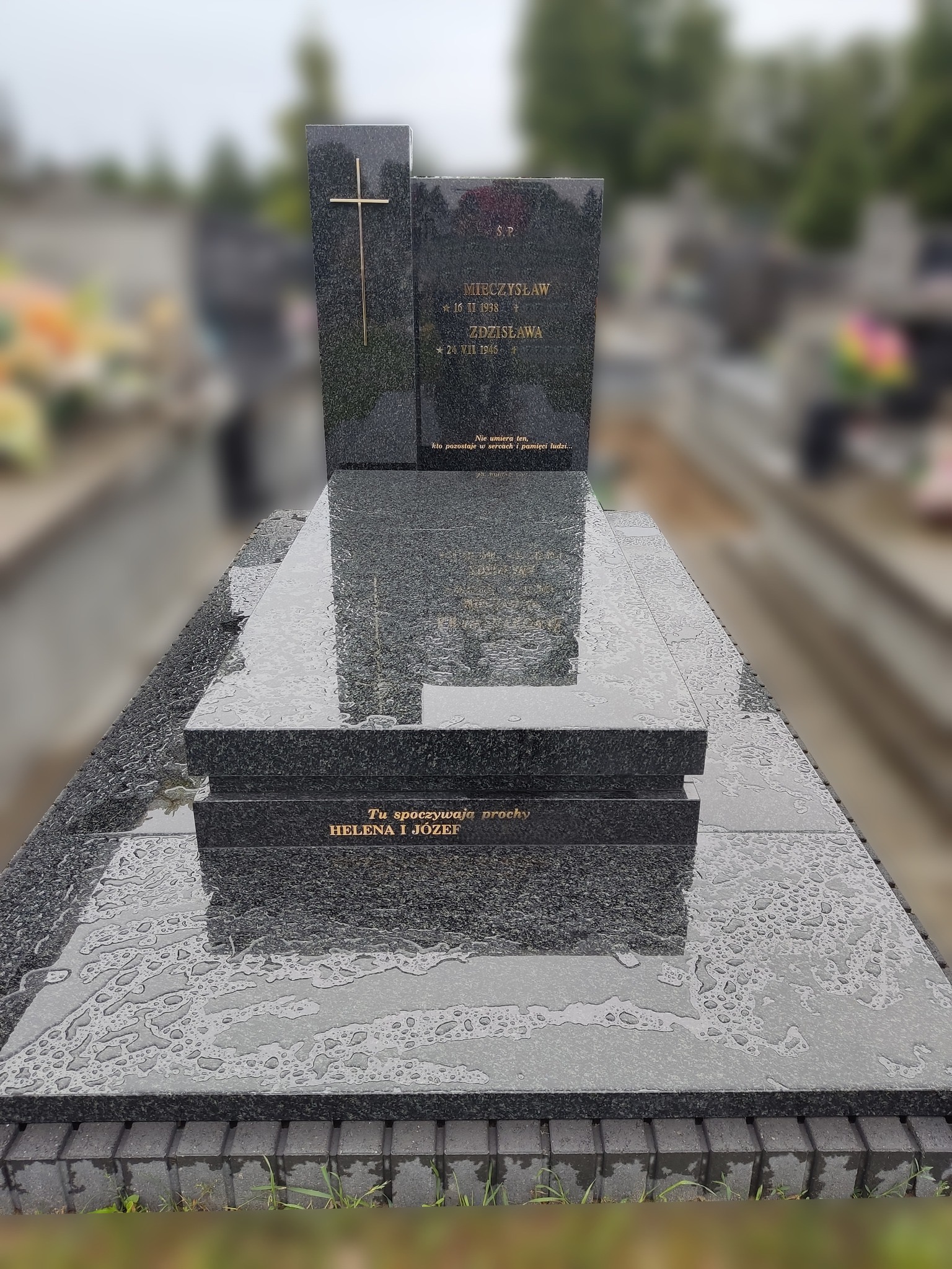Granitowy nagrobek z wygrawerowanym krzyżem i inskrypcjami, mokry od deszczu, na cmentarzu z rozmytym tłem.