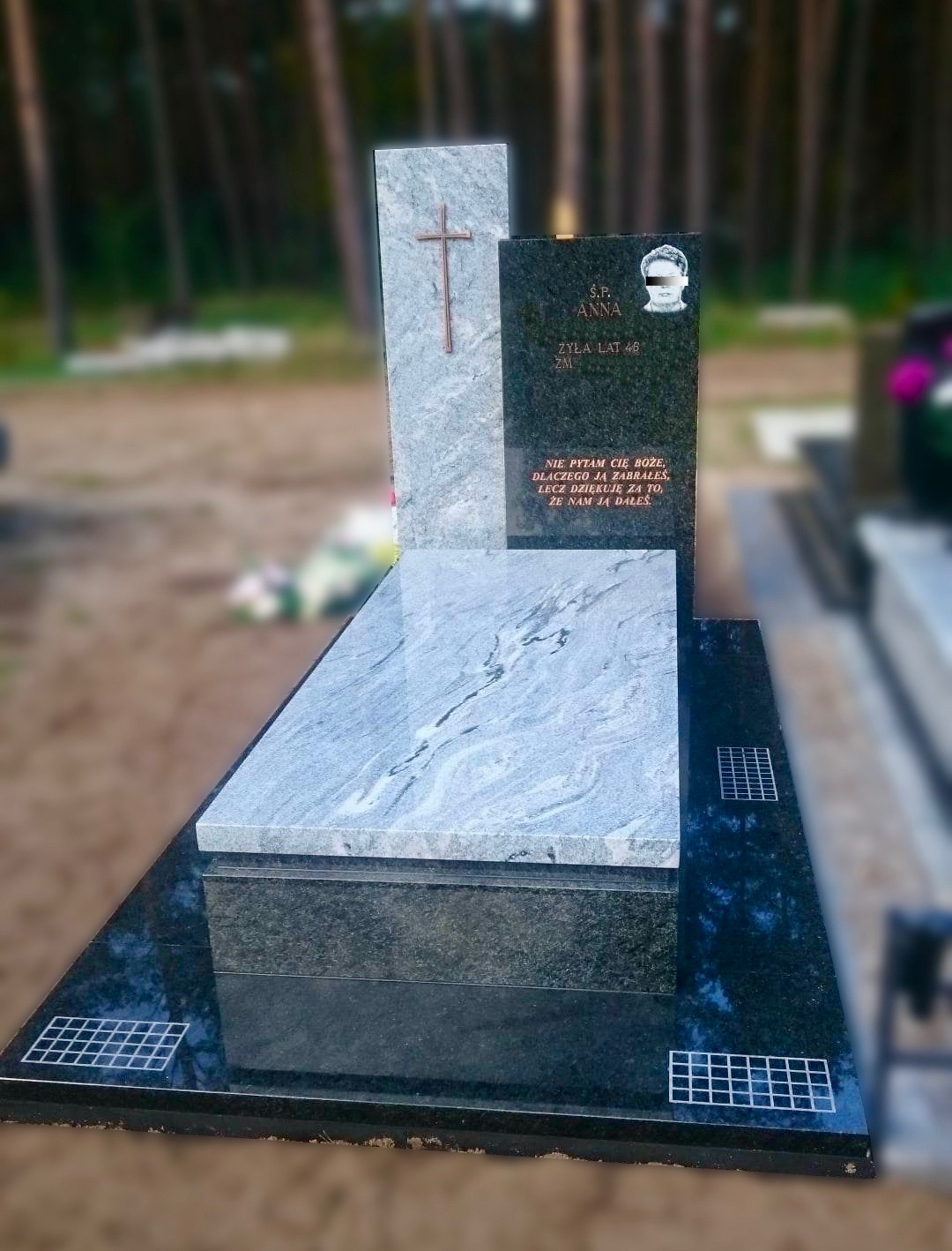 Granitowy pomnik z krzyżem i wygrawerowanym portretem na czarnej tablicy, otoczony zielenią drzew.