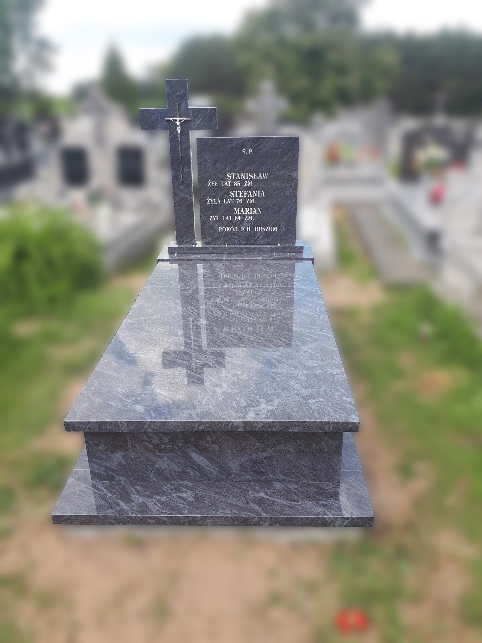 Szary, dwupoziomowy nagrobek z granitu z krzyżem i inskrypcjami, na tle cmentarza