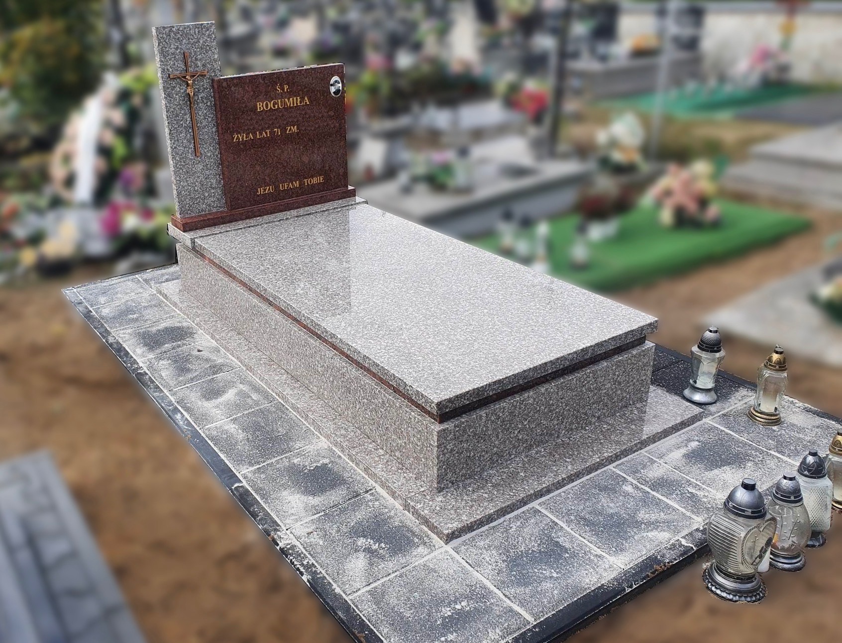 Granitowy pomnik z ciemnobrązową tablicą inskrypcyjną i krzyżem, otoczony kostką brukową, na tle cmentarza.