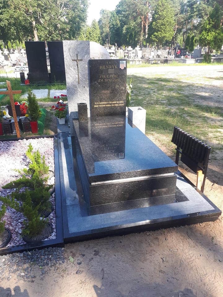 Czarny, polerowany nagrobek z inskrypcją 'Henryk' i datą śmierci, otoczony zielenią cmentarza i drobnymi roślinami w doniczkach.