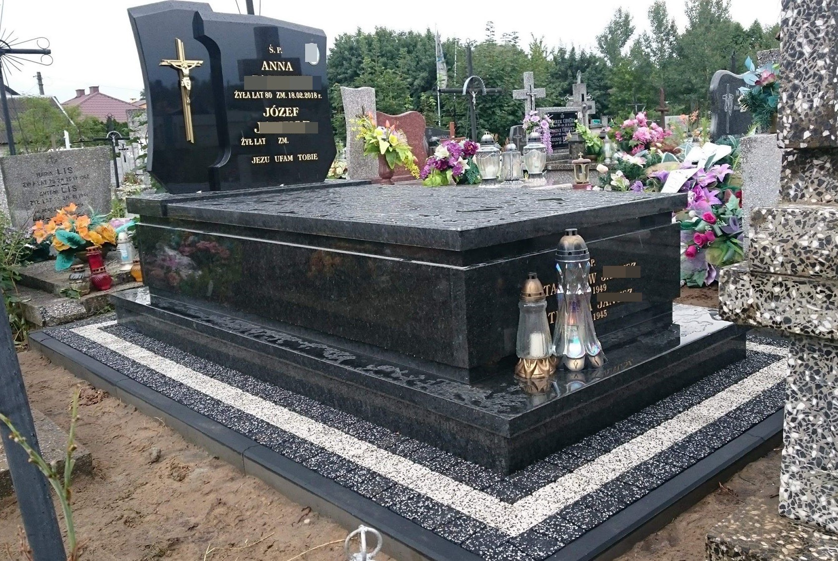 Czarny, granitowy nagrobek z krzyżem i inskrypcjami, otoczony dekoracyjną obwódką z kruszywa w dwóch kolorach, z zapalonymi zniczami i kwiatami w tle.